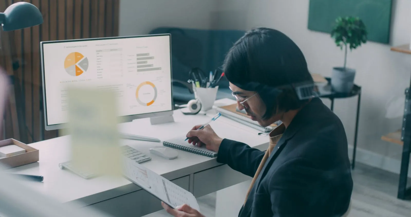 Pessoa anota num caderno em frente a um monitor com gráficos de um relatório financeiro
