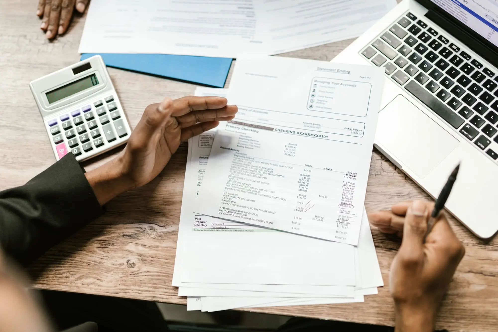 Mãos conferindo um extrato bancário impresso, com calculadora e notebook na mesa
