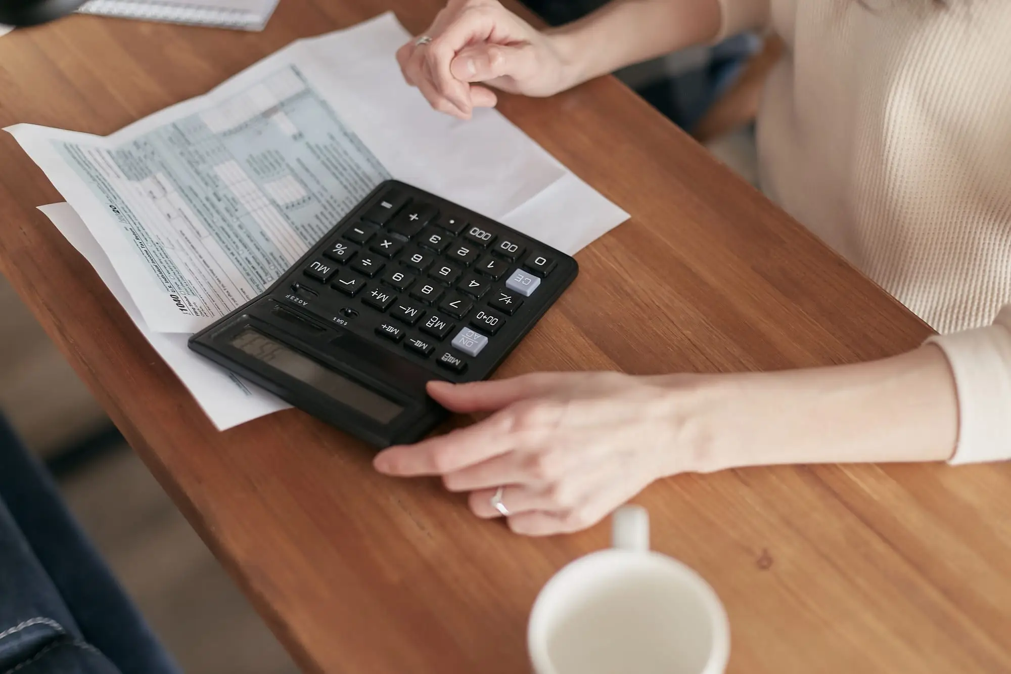 Mãos sobre uma calculadora ao lado de contas impressas, numa mesa de madeira
